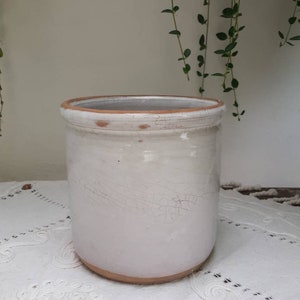 May include: White ceramic planter with a distressed finish and a brown rim. The planter is sitting on a white lace tablecloth.