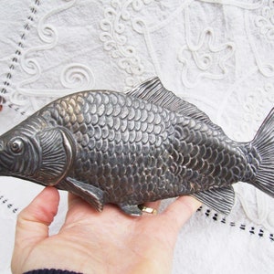 Könnte beinhalten: Ein silberfarbenes, fischförmiges Dekorationsobjekt. Der Fisch hat detaillierte Schuppen, Flossen und einen Schwanz. Das Objekt wird in einer Hand gehalten, der Hintergrund ist ein weißes, gemustertes Gewebe.