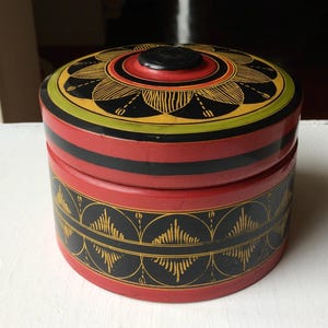 May include: A round, lidded wooden box featuring a vibrant red and black colour scheme. The lid displays a central black circular knob surrounded by a yellow and black floral pattern. The sides are decorated with a repeating black diamond motif.