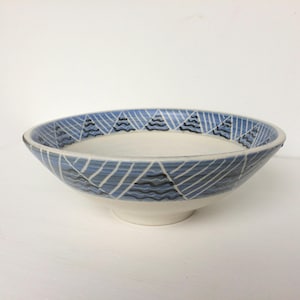 May include: A shallow ceramic bowl with a white interior and a light blue exterior featuring a repeating pattern of dark blue wavy lines inside triangular shapes outlined in white. The bowl has a wide rim and a circular base, showcasing a hand-painted aesthetic.