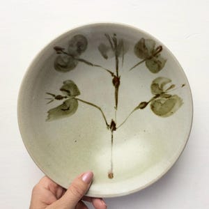 May include: A round ceramic bowl with a light beige, speckled glaze. The interior features a hand-painted botanical design in muted olive green and dark brown, depicting a central stem with four delicate, abstract flower blossoms.