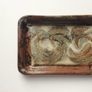 May include: Rectangular ceramic tray with rounded corners. The tray has a textured surface with a brown, tan, and blue swirl design. The edges are a darker brown, giving the tray a rustic appearance.