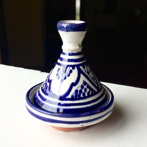 May include: A blue and white ceramic tagine pot. The conical lid and round base are decorated with blue patterns and stripes on a white background. The base is reddish-brown. A traditional cooking pot.
