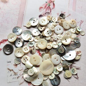 May include: A large collection of assorted round buttons in various sizes and shades of white, cream, grey, and iridescent pearl. The buttons feature two or four holes and exhibit smooth, glossy, or textured surfaces with varying levels of lustre.