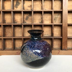 May include: A small, round ceramic vase with a narrow neck and a flared rim. The surface features a mottled glaze with deep shades of navy blue, purple, and black, transitioning into a textured, speckled light grey pattern on the lower section.