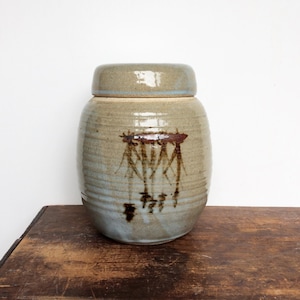 May include: A cylindrical ceramic jar with a matching lid, featuring a light beige, speckled glaze with a smooth, glossy finish. The centre of the jar displays a dark brown, abstract, brush-stroke design that resembles hanging roots or branches.