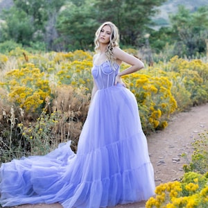 May include: A woman in a lavender tulle gown with a fitted bodice and a flowing skirt stands in a field of yellow wildflowers. The gown has a sweetheart neckline and a fitted bodice with a corset-like design. The skirt is full and flowing, with multiple layers of tulle. The woman is looking at the camera with a serious expression.