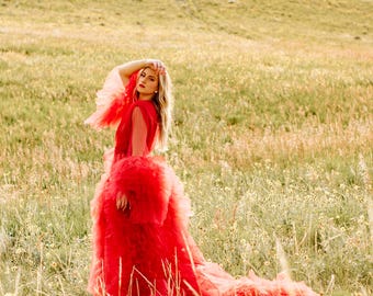 May include: A woman in a bright red tulle dress poses in a field of green grass and yellow wildflowers.