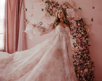 May include: A woman wearing a long, flowing, pink floral print dress with large, puffed sleeves. She is standing in front of a pink wall with a floral backdrop.