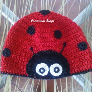 Può includere: Cappello di coccinella in crochet rosso con punti neri e un viso nero con occhi bianchi. Il cappello ha un bordo in crochet nero attorno al bordo. Il testo "Creazioni Fiopi" è visibile sul cappello.