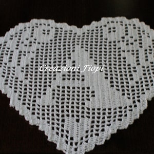 Può includere: Un centrino a forma di cuore in pizzo bianco lavorato a uncinetto con un delicato motivo floreale. Il centrino è realizzato con un filo sottile e ha un aspetto delicato e traforato. Il testo "Creazioni Fiopi" è visibile al centro del centrino.