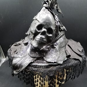 May include: A black hat featuring a molded skull centerpiece integrated into the crown. The hat is constructed from layered leather-like material with a wide brim edged in intricate black lace trim and dangling teardrop-shaped fringe accents.