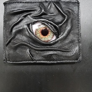 May include: A rectangular black leather wallet featuring a textured, wrinkled surface that forms a raised, organic shape around a central, realistic graphic of a human eye with a green iris and a dark pupil, stitched along the edges.