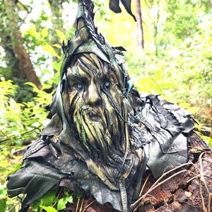 May include: A sculpted mask featuring a weathered, tree-like face with deep grooves, a pointed hood, and layered leather-like textures in shades of forest green, bronze, and black. The figure has piercing light-colored eyes and a long, textured beard.
