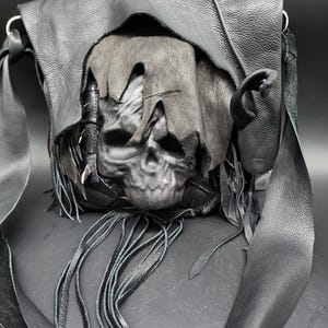 Puede incluir: Bolso de hombro de cuero negro con un diseño de calavera esculpida. El bolso tiene una solapa que cubre la calavera, flecos y correas largas. El bolso está hecho de cuero texturizado y tiene herrajes plateados.