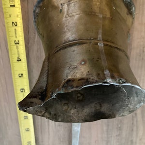 May include: A vintage brass bell with a flared bottom and a long, thin handle. The bell has a tarnished, aged appearance with visible wear and tear. The bell's height is approximately 4 inches.