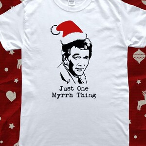 May include: White t-shirt featuring a black and white portrait of a man wearing a red Santa hat. The text "Just One Myrrh Thing" is printed below the portrait in black. The shirt is a festive holiday design.