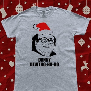 Può includere: Una maglietta grigia con un'illustrazione in bianco e nero di Danny DeVito che indossa un cappello da Babbo Natale. Il testo "DANNY DEVITHO-HO-HO" è stampato sotto l'illustrazione.