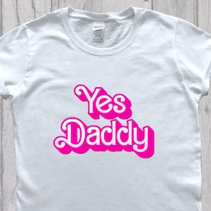 Pode incluir: Camiseta branca com o texto "Yes Daddy" em rosa, em letras de estilo retrô.
