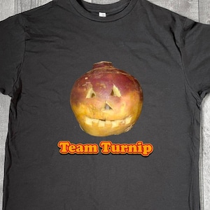 May include: Dark gray t-shirt featuring a carved turnip with a smiling face. The text "Team Turnip" is printed in orange and yellow below the turnip. The shirt is made of a soft material.