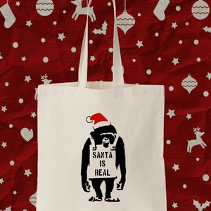 May include: A white canvas tote bag featuring a black and red image of a gorilla wearing a Santa hat and holding a sign that reads "Santa is Real".