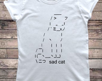 Può includere: Maglietta bianca con un design di gatto triste in ASCII nero. Il testo "sad cat" è sotto il gatto.