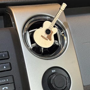 May include: A wooden guitar-shaped car air vent clip. The guitar has a light brown body with a darker brown soundhole and fretboard. It is inserted into a silver car air vent, with a black control panel and a 12V power outlet visible.