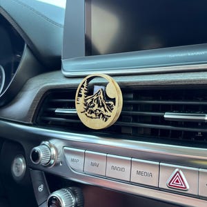 May include: A round, wooden car air vent diffuser with a mountain and tree design. The diffuser is made of light-colored wood and is placed in a car's air vent. The design features a mountain range, trees, and a crescent moon.