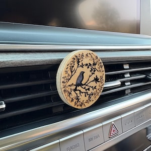 May include: A round, wooden car air freshener featuring a laser-etched design of a bird perched on a branch within a circular frame of leaves. The bird is a muted blue-gray color. The freshener is attached to a car vent.