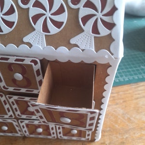 May include: A cardboard advent calendar shaped like a gingerbread house with numbered drawers. The drawers are decorated with white and red candy cane designs. The top of the house is decorated with white and red candy cane designs.