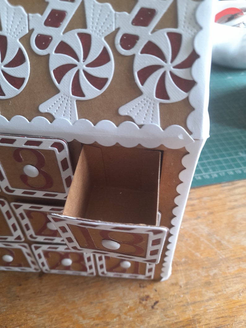 May include: A cardboard advent calendar shaped like a gingerbread house with numbered drawers. The drawers are decorated with white and red candy cane designs. The top of the house is decorated with white and red candy cane designs.