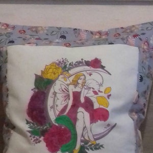 May include: Cushion with a fairy and floral design. The central image shows a woman in a red dress on a crescent moon, surrounded by pink, yellow, and purple flowers. The border has a grey floral pattern.