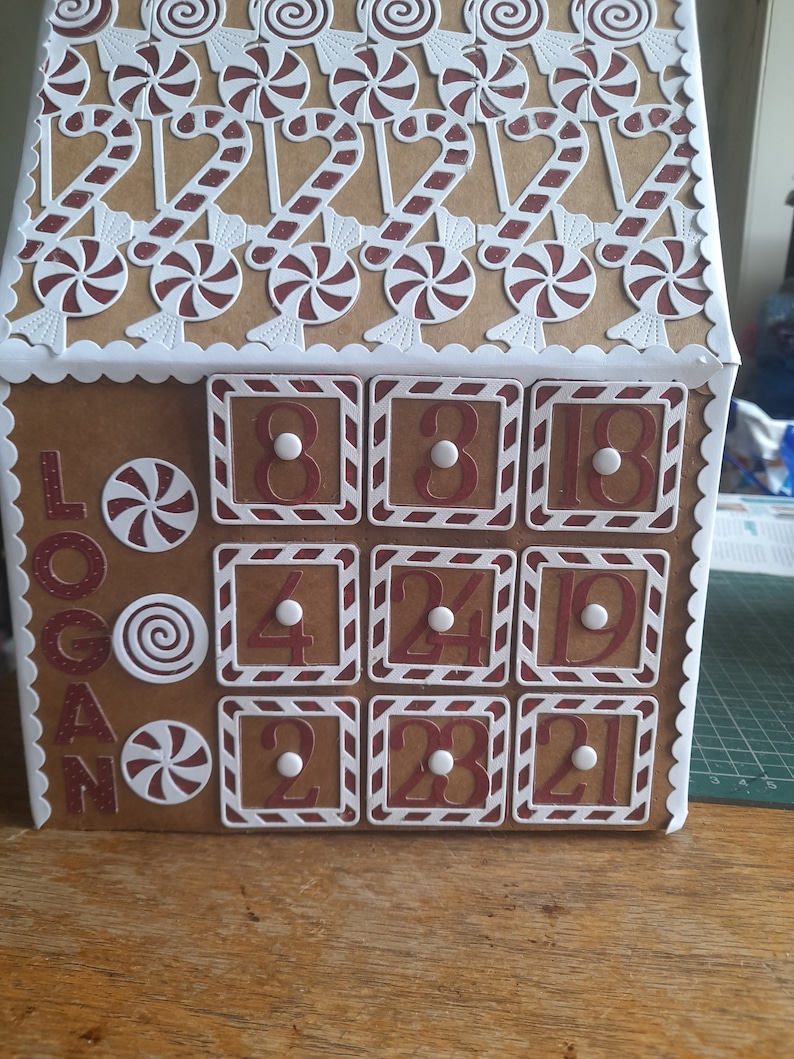 May include: A cardboard gingerbread house advent calendar with red and white candy cane decorations and numbered doors. The doors are labelled with the numbers 8, 3, 18, 4, 24, 19, 2, 23, and 21.