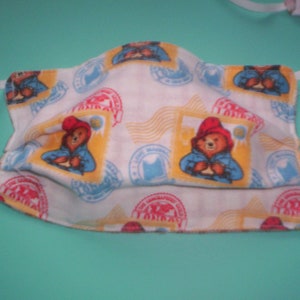 Peut inclure: Masque facial en tissu blanc avec un motif r&eacute;p&eacute;t&eacute; d'un ours portant un chapeau rouge et une chemise bleue. L'ours tient un colis. Le fond est blanc avec des accents jaunes et bleus.