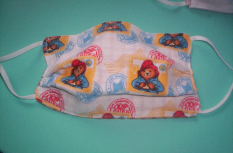 Peut inclure: Masque facial en tissu blanc avec un motif r&eacute;p&eacute;t&eacute; d'un ours portant un chapeau rouge et une chemise bleue. L'ours tient un colis. Le fond est blanc avec des accents jaunes et bleus.