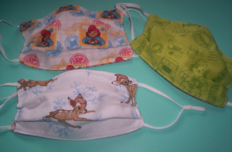 Peut inclure: Trois masques en tissu avec des motifs diff&eacute;rents. Un masque a un fond jaune avec un motif r&eacute;p&eacute;t&eacute; de timbres rouges et bleus. Un autre masque a un fond vert avec un motif r&eacute;p&eacute;t&eacute; de timbres verts et blancs. Le troisi&egrave;me masque a un fond blanc avec un motif r&eacute;p&eacute;t&eacute; de flocons de neige bleus et un cerf brun.
