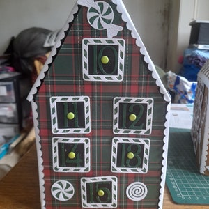 May include: A red and green tartan advent calendar shaped like a house with white scalloped edges. The numbers 10, 11, 12, 13, 14, and 15 are visible on the front of the calendar. Each number is surrounded by a white frame with black accents. There are two white candy cane decorations on the front of the calendar.