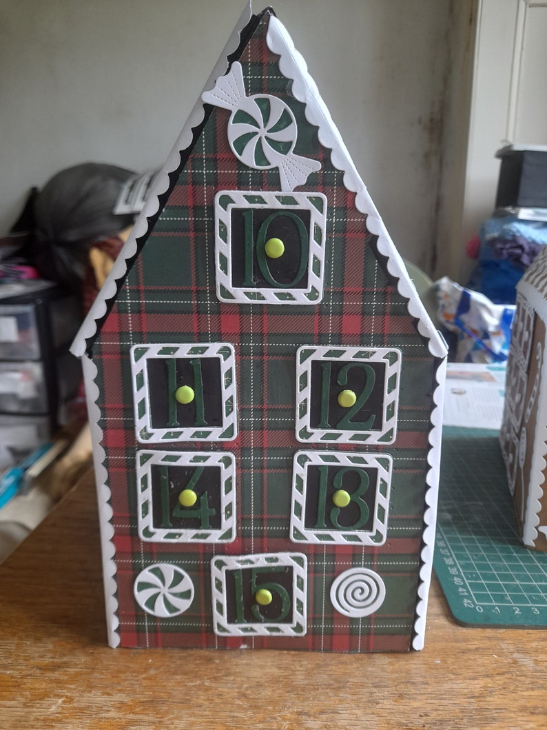 May include: A red and green tartan advent calendar shaped like a house with white scalloped edges. The numbers 10, 11, 12, 13, 14, and 15 are visible on the front of the calendar. Each number is surrounded by a white frame with black accents. There are two white candy cane decorations on the front of the calendar.