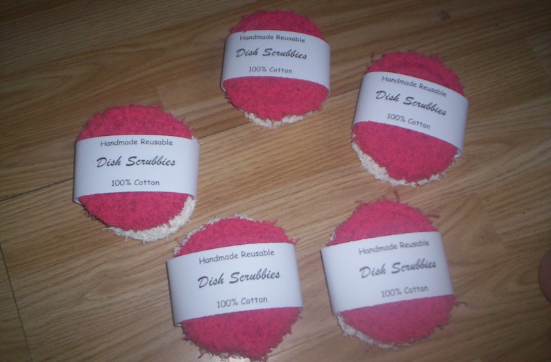 May include: Five pink and white handmade reusable dish scrubbies with a white label that reads "Handmade Reusable Dish Scrubbies 100% Cotton".