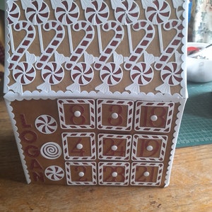 May include: A brown cardboard advent calendar shaped like a house with white trim and candy cane details. The front of the calendar has 12 numbered doors with white trim and red candy cane details. The calendar is decorated with white paper candy canes and lollipops. The text "Logan" is written on the side of the calendar.