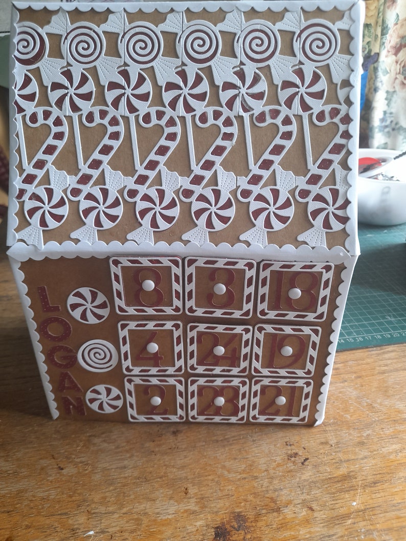 May include: A brown cardboard advent calendar shaped like a house with white trim and candy cane details. The front of the calendar has 12 numbered doors with white trim and red candy cane details. The calendar is decorated with white paper candy canes and lollipops. The text "Logan" is written on the side of the calendar.