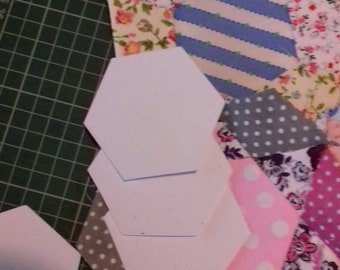 May include: A close-up of a green cutting mat with several white hexagon-shaped fabric pieces and a patchwork of colourful fabric scraps. The fabric scraps are in various patterns, including floral, polka dot and striped.