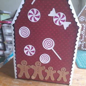 May include: A red gingerbread house with white trim and a white scalloped edge. The house is decorated with pink and white candy canes, swirls, and a bow. There are five gingerbread men at the bottom of the house.