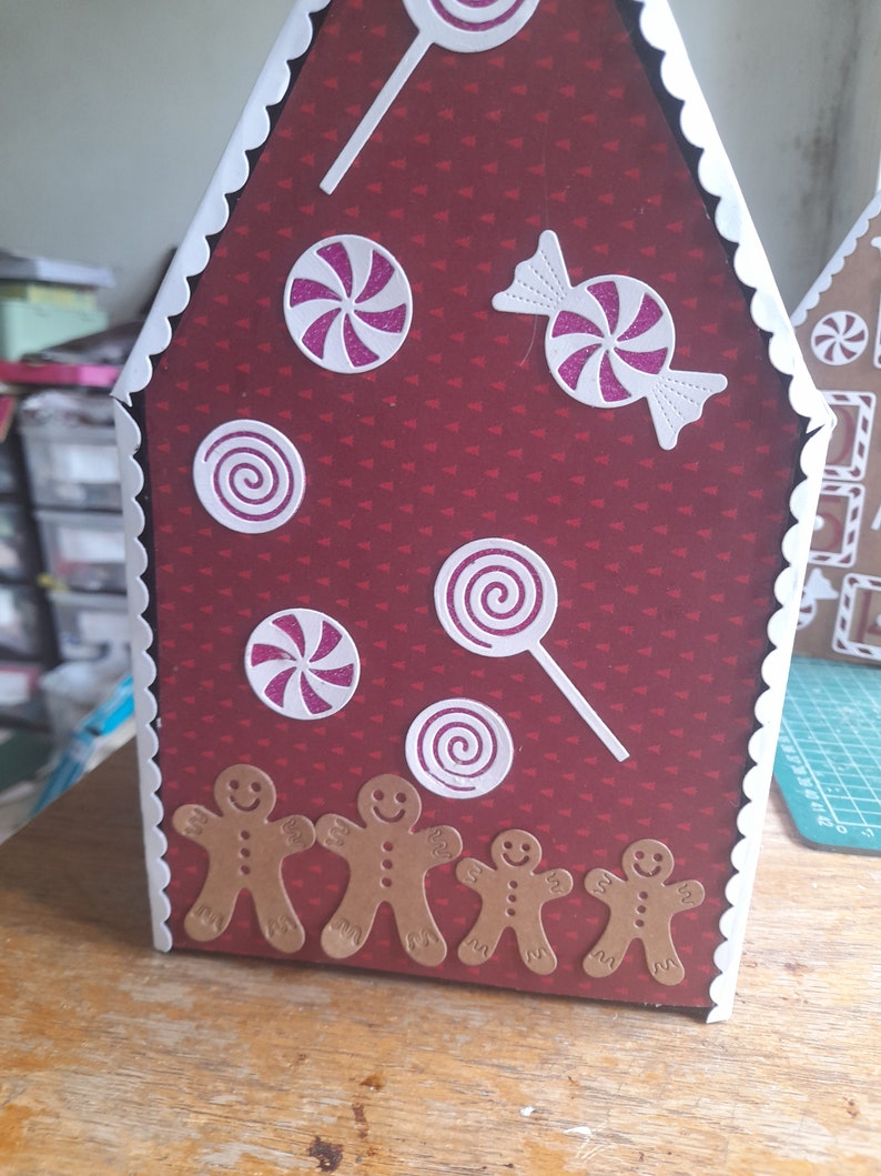 May include: A red gingerbread house with white trim and a white scalloped edge. The house is decorated with pink and white candy canes, swirls, and a bow. There are five gingerbread men at the bottom of the house.