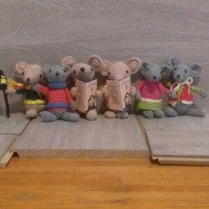 May include: Seven grey crocheted mice holding small books. The mice are wearing colourful sweaters and are standing on a wooden surface.
