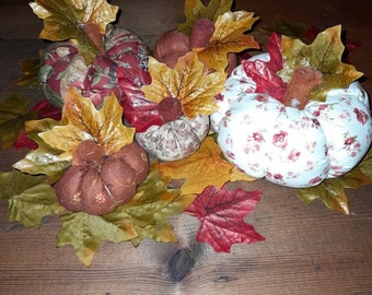 May include: Four fabric pumpkins with autumn leaves. The pumpkins are in various patterns, including floral and plaid. The leaves are in shades of red, orange, and yellow.