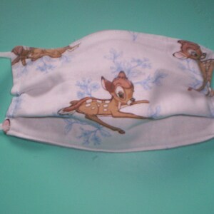 Peut inclure: Masque facial blanc avec un motif de flocons de neige bleu et blanc et une image r&eacute;p&eacute;t&eacute;e de Bambi le cerf. Le masque a des lani&egrave;res &eacute;lastiques blanches.