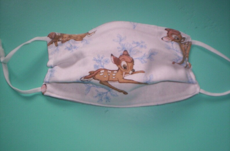 Peut inclure: Masque facial blanc avec un motif de flocons de neige bleu et blanc et une image r&eacute;p&eacute;t&eacute;e de Bambi le cerf. Le masque a des lani&egrave;res &eacute;lastiques blanches.