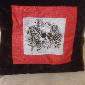 May include: Black pillow with a red border and a white square in the centre. The white square features a black and white illustration of a skull with roses and a butterfly.