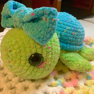 May include: A crocheted turtle toy with a blue bow and green and blue speckled yarn. The turtle has large, black eyes and a pink nose.